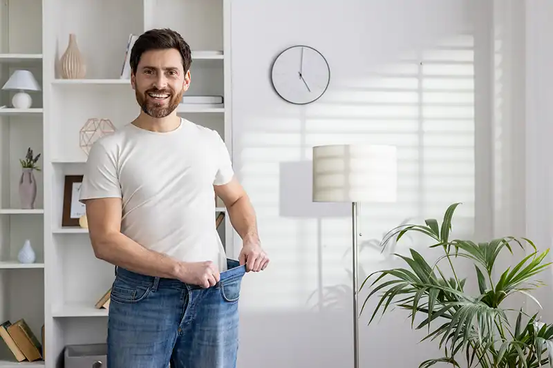 smiling man showing physician supervised weight loss by pulling pants at waist